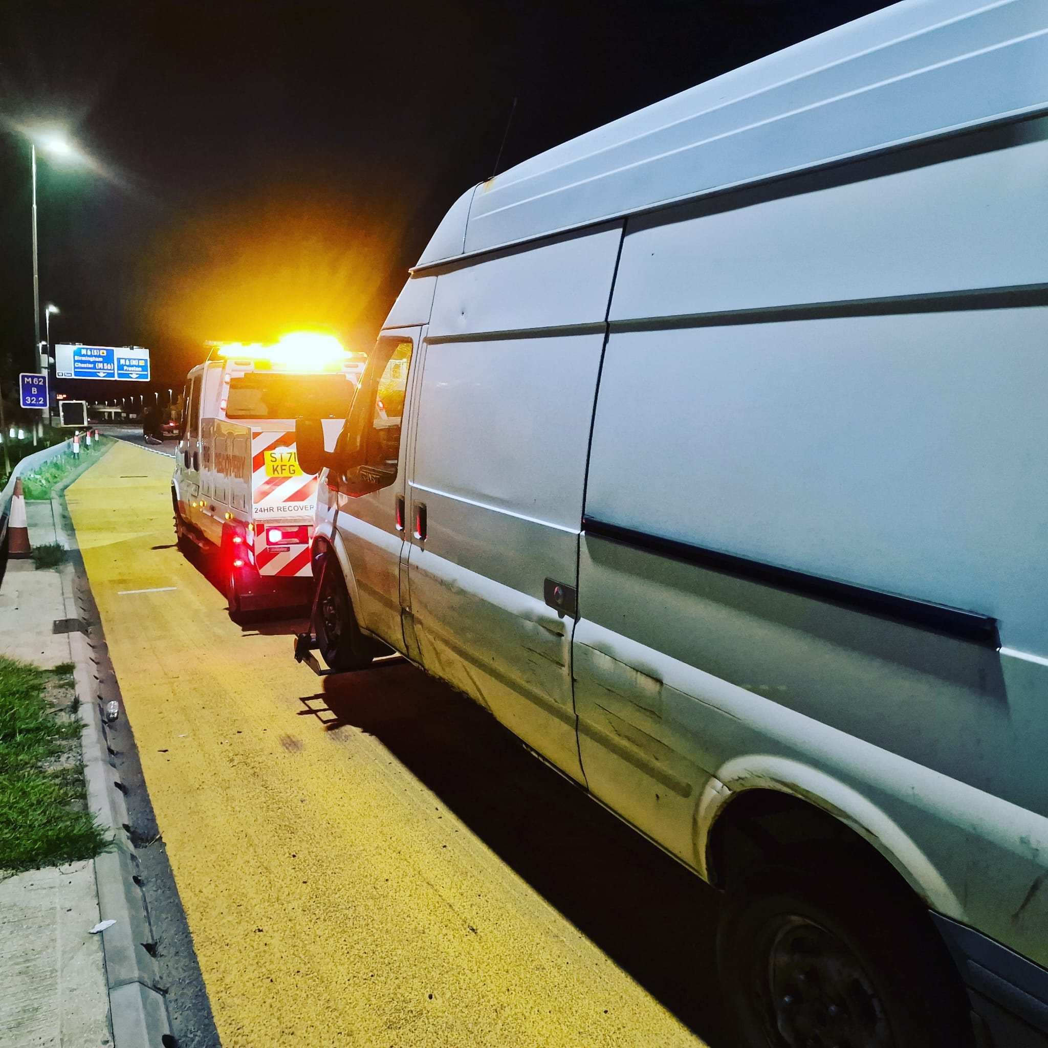 Red DAF LF recovery truck BE61 V25 parked along a coastal road with a pier in the distance, operator in high-vis attending a stranded car ahead