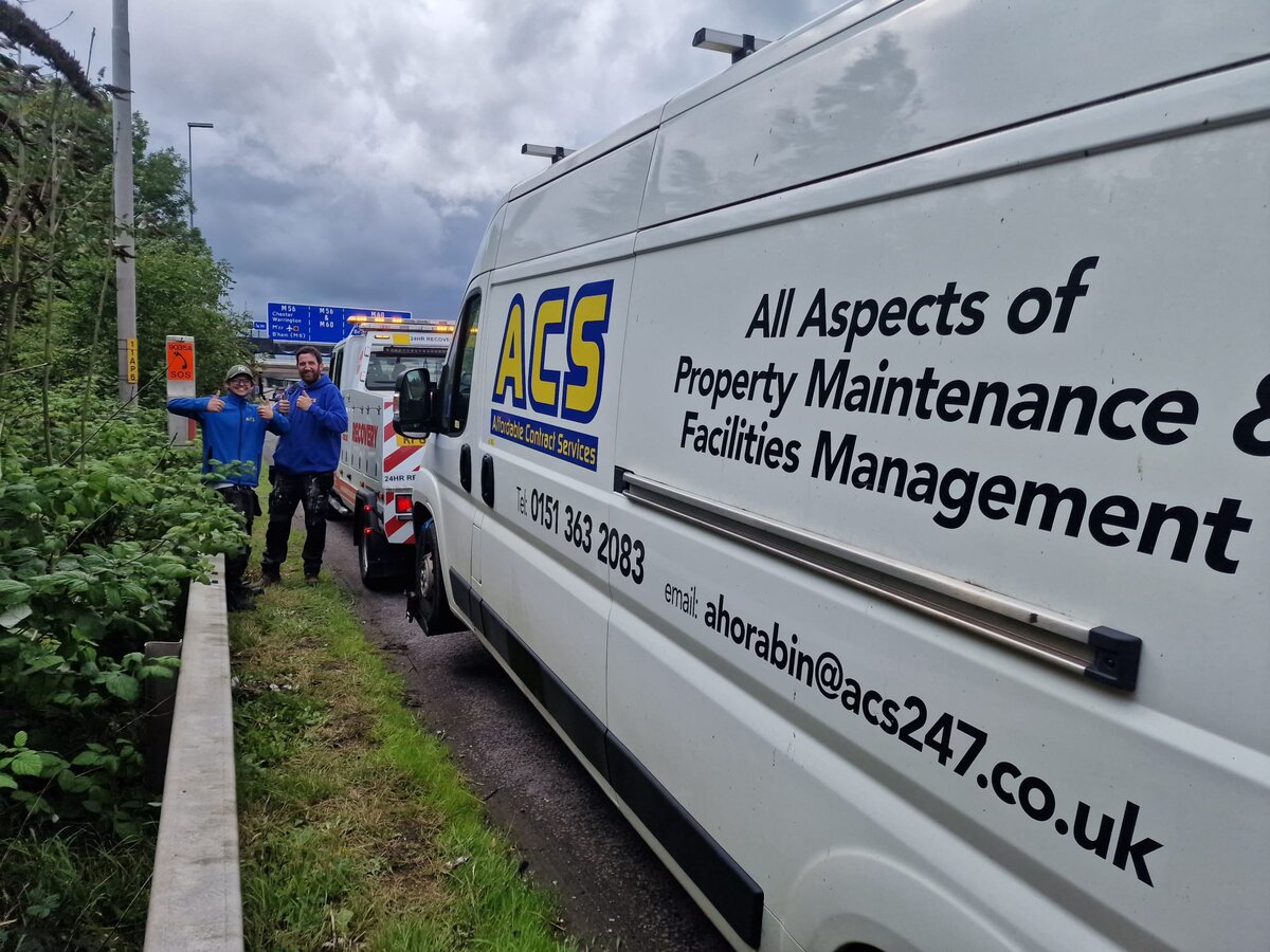 A driver standing beside an ACS Property Maintenance van on a grass roadside verge on the M62, with the Breakdown Man recovery truck positioned behind ready to load.