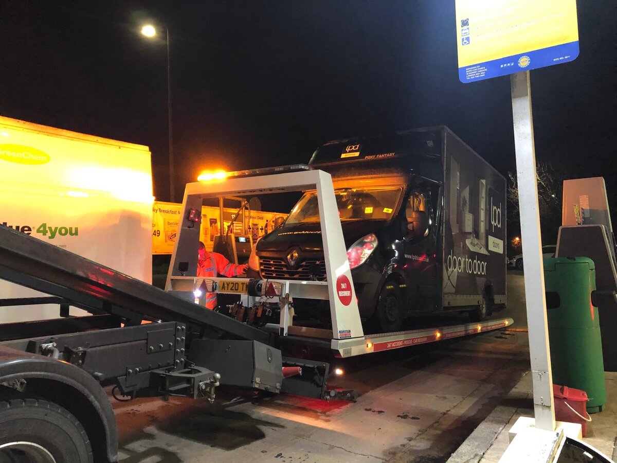 Black LPD door-to-door courier van being loaded onto the Breakdown Man recovery truck under petrol-station forecourt lights at night