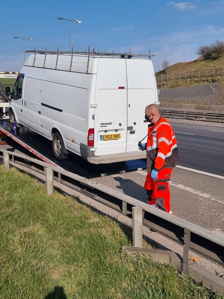 White Ford Transit van loaded on a Breakdown Man recovery truck on a motorway-side verge with the operator in orange hi-vis stood beside it.