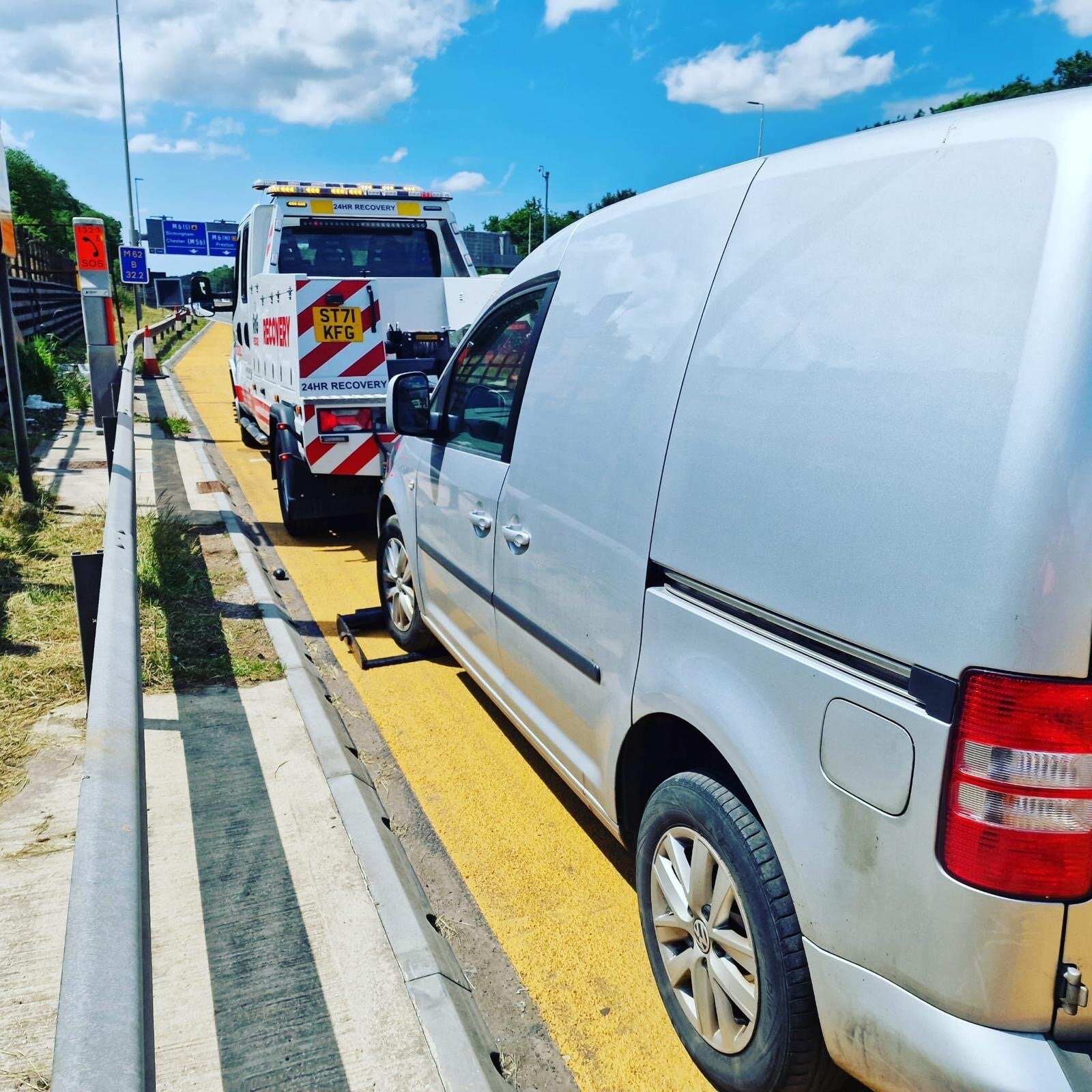 A silver VW Caddy van pulled into a yellow-hatched motorway emergency refuge area with a 24-hour recovery truck positioned in front, ready to load.