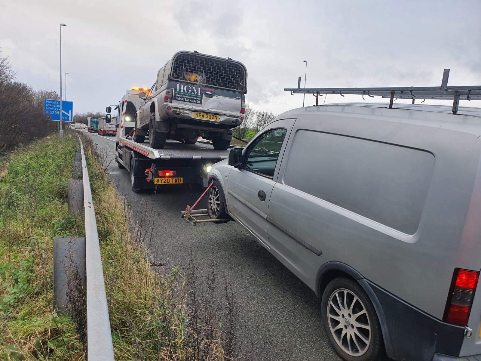 A Land Rover Defender loaded onto a recovery truck on the M6 hard shoulder, alongside a stranded grey van waiting to be cleared.